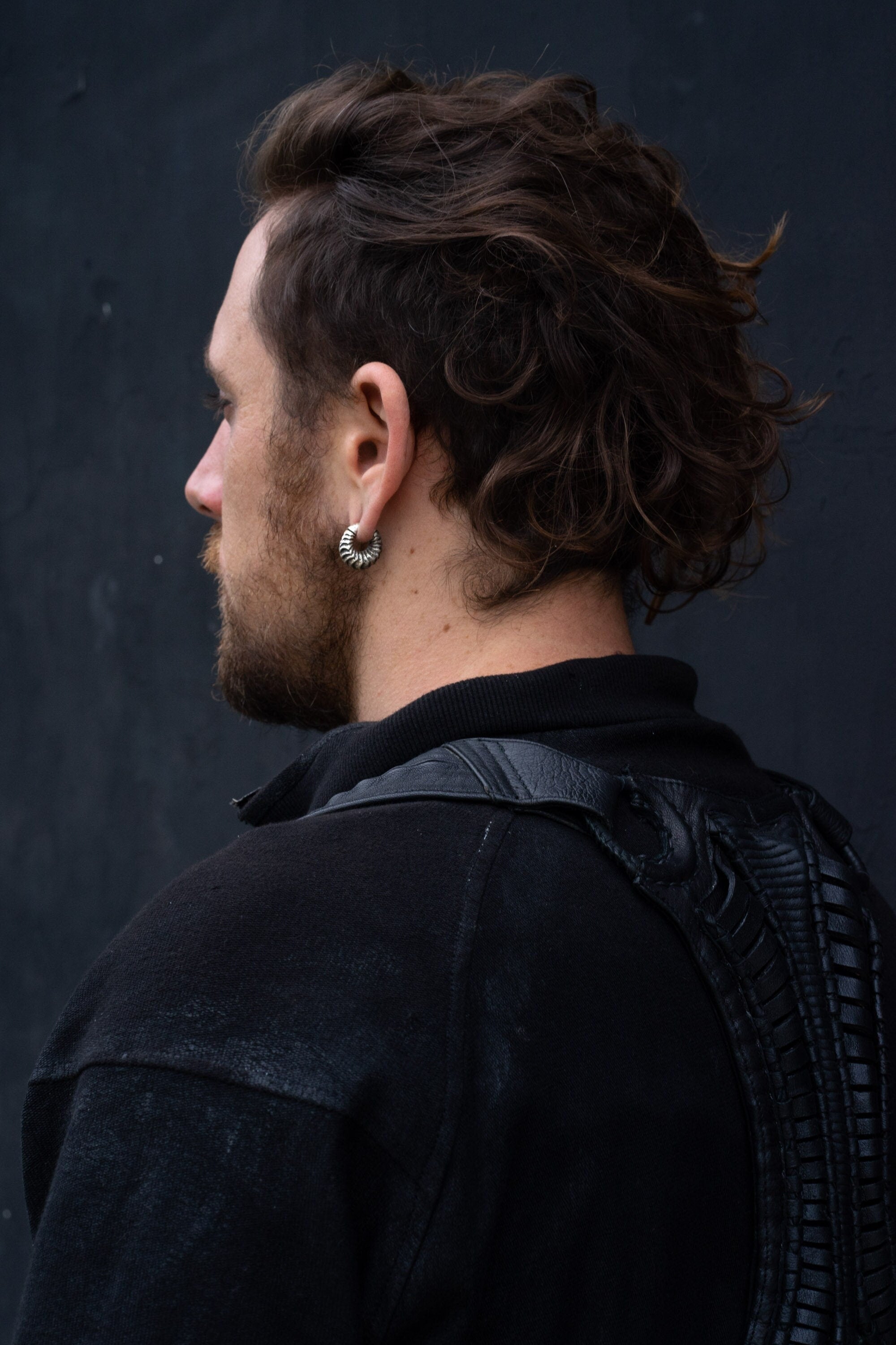 A person with medium-length wavy hair and a beard is seen from the side and back, showcasing their Kaya Movement Fractal Alien Bio-Organic Circle Ear Weights in modern brass 4mm (6ga) gauges designed for stretched lobes. They are wearing a dark jacket featuring a unique textured design on the back against a plain, dark wall background.